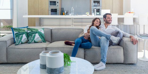 Couple sitting in a modern open plan house. They are sitting on a sofa with a kitchen behind them. They are attractive, smiling and happy.