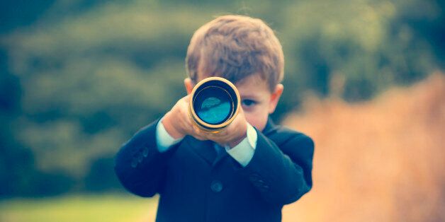 Young boy in a business suit with telescope. Small child wearing a full suit and holding a telescope. He is holding the telescope up to his eye. Business forecasting, innovation, leadership and planning concept. Shot outdoors with trees and grass in the background