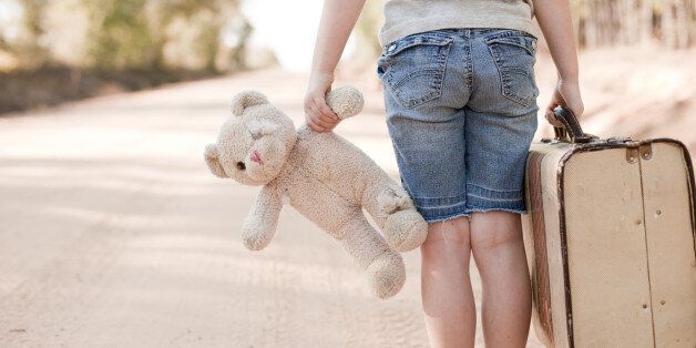 'Color photo of a sad, lonely 6-7 year old girl carrying an old, raggedy teddy bear and an old suitcase while standing on a dirt road outside.'