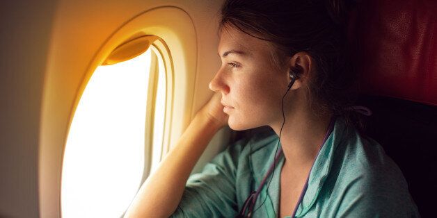 Young woman sitting in the plane, looking away through the window and listeting music with headphones