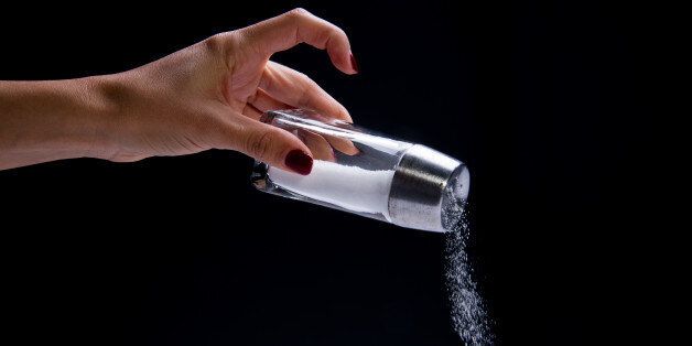 glass salt shaker with brushed steel capA hand is holding a glass and steel salt shaker and pouring out granules of salt, against a black background