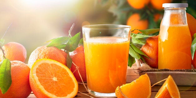 Glass of orange juice on a wooden table with bottle and orange sections. Tree and field background with evening sun. Horizontal composition. Front view