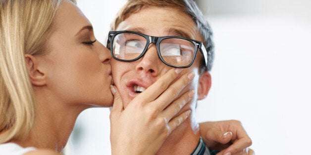 Beautiful blonde kissing male geek on the cheek while male looks alarmed and looks sideways