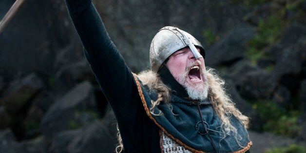 Partial view of a Viking male reenactor with long blond hair under a metal helmet dressed in full warrior armour and battle gear with raised weapon yelling a victory cry in battle in the historic location where Vikings once assembled annually to recite and discuss laws, Pingvellir, Iceland