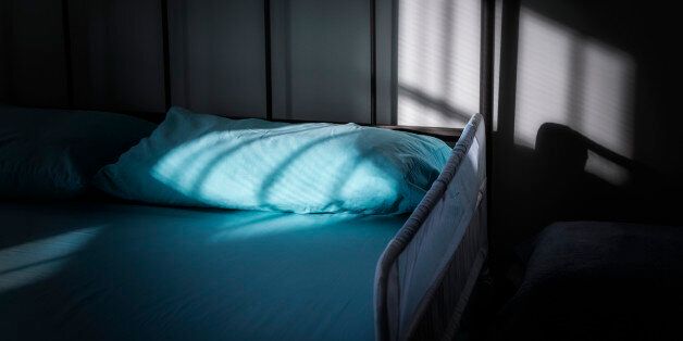 A protective safety side rail guarding the edge of an aging senior adult's bed in a darkened, melancholic bedroom naturally lit by late afternoon sunlight through a venetian blind on an off-camera window. The side rail metal frame and netting helps prevent a restless sleeping person with an illness or other aging or health issues from falling out of bed at night.