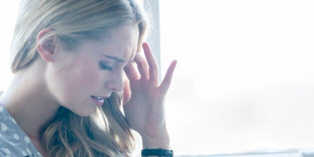 Woman looking upset or in pain. Attractive woman leaning against a wall with her hand on her forehead. She could be in pain, upset, angry, depressed or stressed. She is frowning and standing in front of a window. She is casually dressed. Copy space on right.