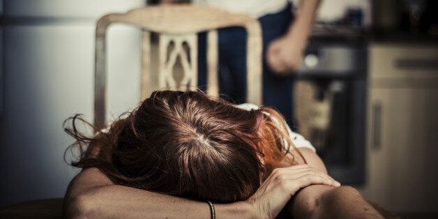 Young woman is reesting her head on a kitchen table, her abusive partner hovering in the background