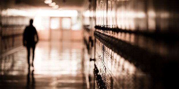 A high school student walks down a dark hallway in a public high school, silhouetted by daylight spilling in and reflecting off of the floor and lockers.