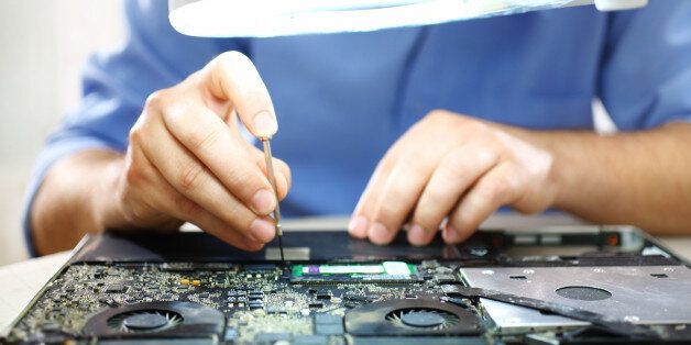 Adult caucasian man unscrewing motherboard on a laptop. He's an employee at computer maintenance and service shop. He's using standing magnifying glass with light, The man is wearing blue shirt with rolled back sleeves.