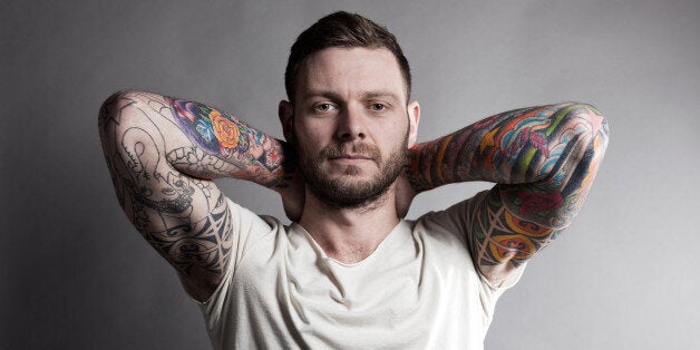 Brunette male, showing tattoo sleeves, with hands on his neck, on a grey background.