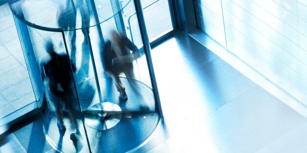 A high angle view of a people entering and exiting an office building through a revolving door. A slow shutter speed allows for the anonymity of the people as they quickly move through the rotating door. A cool blue cast dominates the scene.