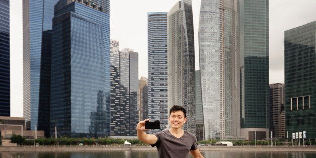 A man in casual clothes takes his own photos against the Singapore skyline with a smart phone