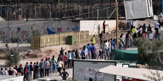 TOPSHOT - Migrants queue next to burned tents at the Moria camp on the Greek island of Lesbos, following the overnight fire on September 20, 2016.Thousands of migrants were forced to flee to safety on September 19, 2016 when their camp on Lesbos was badly damaged in a fire apparently set on purpose, police said. On Lesbos itself there are in excess of 5,600 people, more than 2,000 more than the nominal capacity of the camps. / AFP / LOUISA GOULIAMAKI (Photo credit should read LOUISA GOULIAMAKI/AFP/Getty Images)