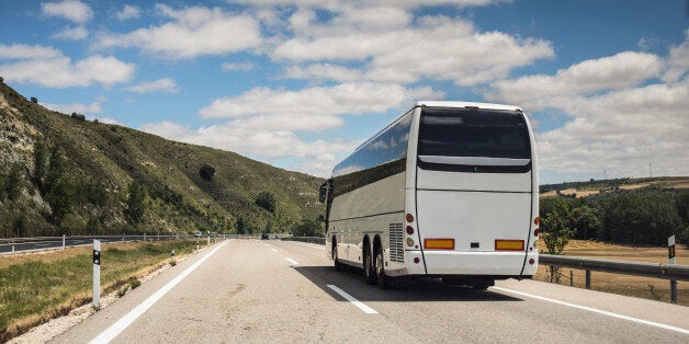 A white coach, or long haul bus for tourists drives through the open roads of Spain, Europe on a summer day. There are white clouds against a blue sky and countryside.