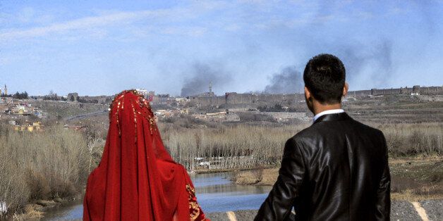 TOPSHOT - A couple hold hands for a wedding photo as they look at smokes rising over the district of Sur in Diyarbakir on February 3, 2016 after clashes between Kurdish rebels and Turkish forces. Four Turkish soldiers were killed on January 27, 2016 in clashes with Kurdistan Worker's Party (PKK) militants in Diyarbakir in southeast Turkey, as a controversial curfew order was expanded to new areas in the Kurdish-dominated city. / AFP / ILYAS AKENGIN (Photo credit should read ILYAS AKENGIN/AFP/Getty Images)
