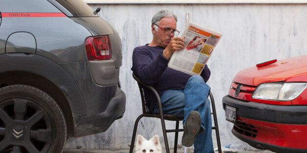 ATHENS, GREECE - APRIL 7: A man reads a newspaper while sitting on a road between two parked cars with his dog, on April 7, 2016 in Athens, Greece. As it recovers from the financial crisis, Athens is experiencing a cultural rebirth. (Photo by Melanie Stetson Freeman/The Christian Science Monitor via Getty Images)