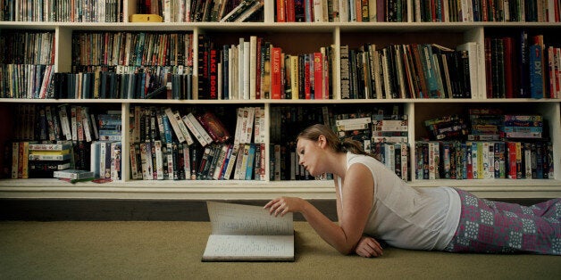 Young woman lying on floor by book shelves, reading, side view