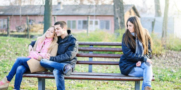 A couple of young man and woman teenagers sitting on a bench, embracing, in love and happy at the other end sits a sad alone girl