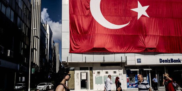 Pedestrians pass a giant Turkish national flag hanging above a DenizBank AS bank branch in Istanbul, Turkey, on Tuesday, July 19, 2016. Turkey's central bank slowed the pace of interest rate cuts at its meeting on Tuesday after the failed coup attempt triggered a sell-off in the currency and sovereign debt. Photographer: Ismail Ferdous/Bloomberg via Getty Images