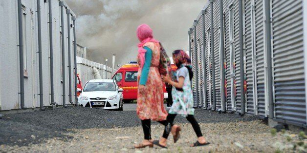Refugees walk past containers as smoke billows in background in the refugee camp near Kassel, Germany, Sunday, Aug. 14, 2016. German authorities say 16 asylum seekers suffered from smoke inhalation after a fire broke out at a container housing facility in the central city of Kassel. The Kassel fire department told the dpa news agency the fire started inside one of the residences Sunday and that an anti-migrant attack had been ruled out. (Bernd Schoelzchen/dpa via AP)