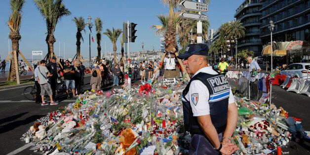 A police officer watches people gathering around a floral tribute for the victims killed during a deadly attack, on the famed Boulevard des Anglais in Nice, southern France, Sunday, July 17, 2016. French authorities detained two more people Sunday in the investigation into the Bastille Day truck attack on the Mediterranean city of Nice that killed at least 84 people, as authorities try to determine whether the slain attacker was a committed religious extremist or just a very angry man. (AP Photo/Laurent Cipriani)