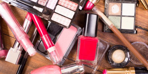 Large pile of various cosmetics lie on top of a wooden dressing table, vanity, or desk. Make-up items include: eye shadow, nail polish, foundation, lipstick, make-up brushes, blush, mascara, lip gloss. Make-up artists' tools. Great background.