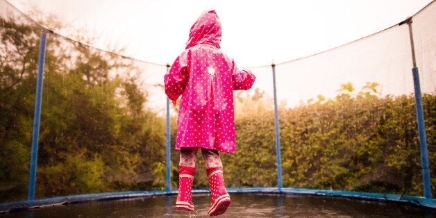 Little child wearing pink raincoat and boots jumping wet trampoline in rain.