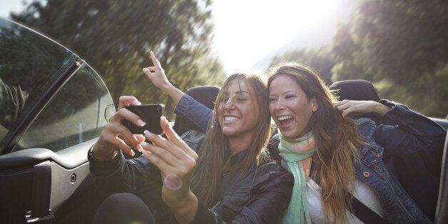 Two friends laughing and making self-portraits with phone. Driving on the backseat of car