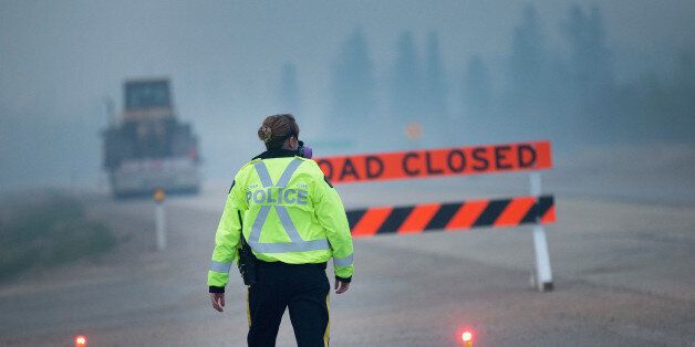 FORT MCMURRAY, AB - MAY 08: Smoke fills the air as a police officer stands guard at a roadblock along Highway 63 leading into Fort McMurray on May 8, 2016 near Fort McMurray, Alberta, Canada. Wildfires, which are still burning out of control, have forced the evacuation of more than 80,000 residents from the town. (Photo by Scott Olson/Getty Images)