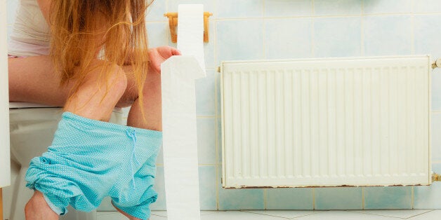 Woman with constipation or diarrhoea sitting on toilet with her blue pajamas down around her legs, holding toilet paper ready in her hands