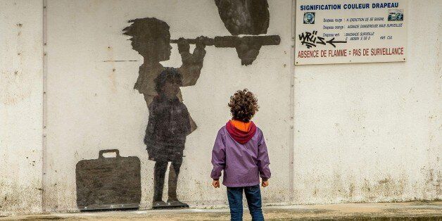 A child looks at an art piece protected by a plexiglass pane by British artist Banksy representing a child with a suitcase looking through a telescope with a vulture perched on it, in tribute to migrants and refugees on December 29, 2015 on a beach in Calais. / AFP / PHILIPPE HUGUEN / RESTRICTED TO EDITORIAL USE - MANDATORY MENTION OF THE ARTIST UPON PUBLICATION - TO ILLUSTRATE THE EVENT AS SPECIFIED IN THE CAPTION (Photo credit should read PHILIPPE HUGUEN/AFP/Getty Images)