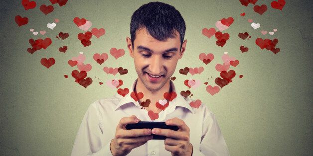 Portrait young handsome happy man sending receiving love sms text message on mobile phone with red hearts flying away up isolated on grey wall background. Human emotions