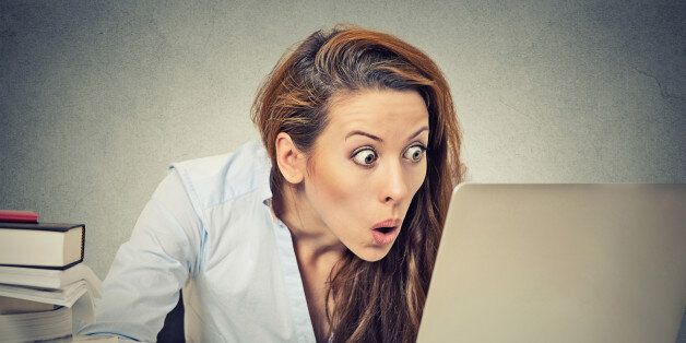 Portrait young shocked business woman sitting in front of laptop computer looking at screen isolated on grey wall background. Funny face expression emotion feelings problem perception reaction