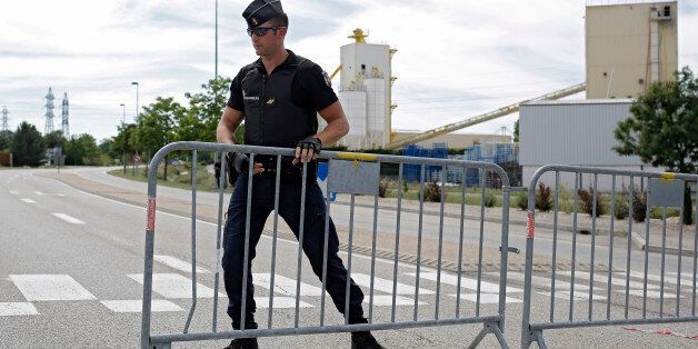 A police officer blocks the road leading to a plant where an attack took place, Friday, June 26, 2015 in Saint-Quentin-Fallavier, southeast of Lyon, France. A man with suspected ties to French Islamic radicals rammed a car Friday into an American gas factory in southeastern France, triggering an explosion that injured two people, officials said. The severed head of a local businessman was left hanging at the factory's entrance, along with banners with Arabic inscriptions, they said. (AP Photo/Laurent Cipriani)