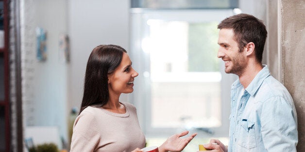 Two cheerful young people holding coffee cups and talking while standing in office