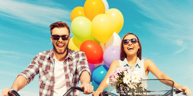 Low angle view of cheerful young couple smiling and riding on bicycles with colorful balloons in the background