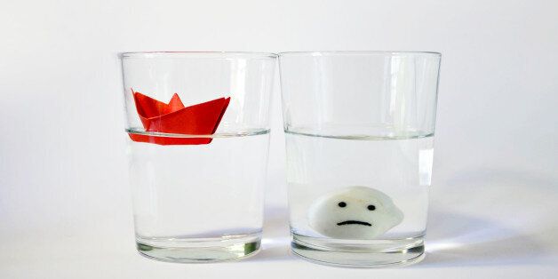 Two glasses of water half full representing two opposite attitudes: optimistic versus pesimistic. The optimistic attitude is represented by a red paper boat floating in the water of the glass. The pesimistic attitude is represented by a white stone with a sad smile on it at the bottom of the other glass.