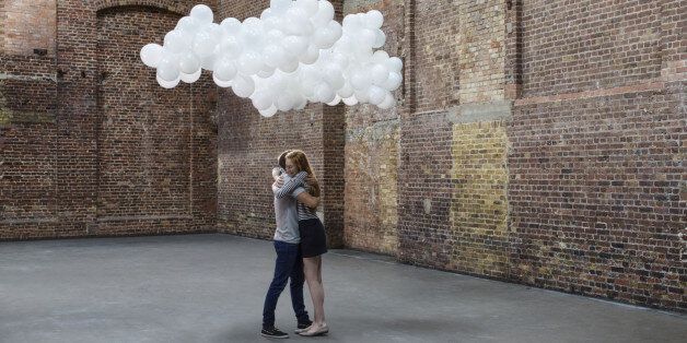 Couple hugging in empty warehouse with cloud made of balloons above heads