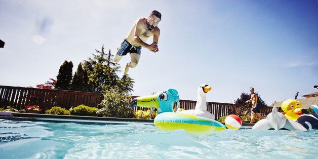 Man diving from pool deck towards inflatable pool toy in outdoor pool with friend watching in background