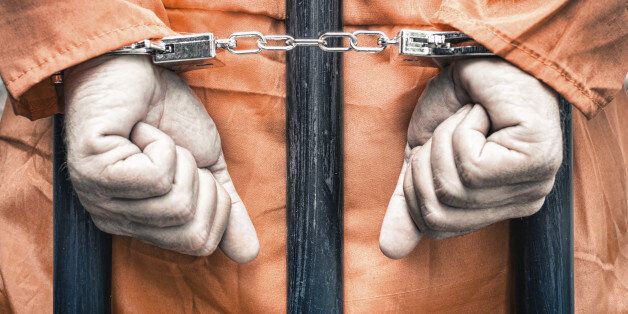 Handcuffed hands of a prisoner behind the bars of a prison with orange clothes - Crispy desaturated dramatic filtered look
