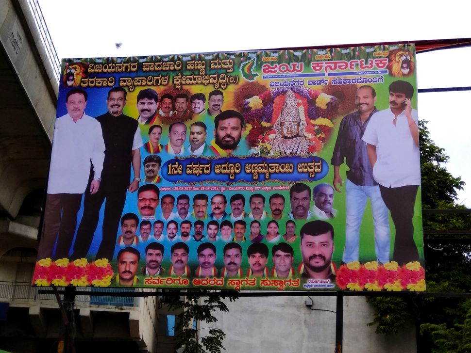 A banner announcing the festival for goddess Annamma features images of MLA Krishnappa and Muthappa Rai, founder of Jaya Karnataka, denoting an equivalence of stature