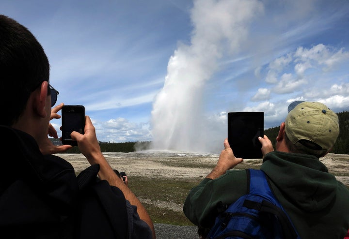 People use phones and tablets to photograph Old Faithful geyser erupting in Yellowstone National Park. The geyser's stea