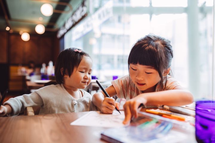 Little girl teaching her little toddler sister to draw joyfully in a restaurant.（イメージ写真）