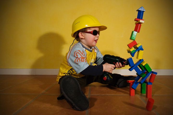 Stop motion shot of a toddler with a yellow helmet, sunglasses and an electric drill having fun in the demolition of his creations made of building bricks.
