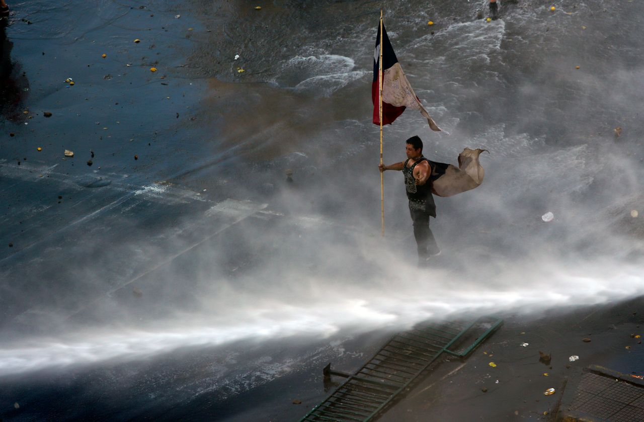 Διαδηλωτής κρατά τη σημαία της Χιλής ενώ δέχεται επίθεση από την αστυνομία με τη χρήση αντλιών νερού. 