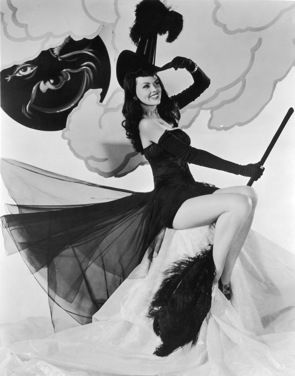 Actress Dusty Anderson poses in a witch's hat and a strapless chiffon costume while holding a feathered broom for Columbia Pictures in the 1940s.