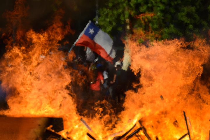 Seen through a burning street barricade, an anti-government demonstrator waves a Chilean flag in Santiago, Chile, on Oct. 28, 2019.