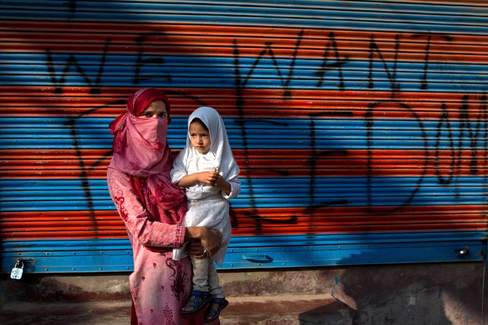 In this Sept. 27, 2019, photo, a Kashmiri woman protestor Jawahira Banoo carries her 3-year-old daughter Rutba and stands for a photograph outside a closed shop with a spray-painted graffiti after a protest on the outskirts of Srinagar. Banoo says she does not miss an opportunity to come out to the streets to protest. The men are at a higher risk of being detained, she says. 