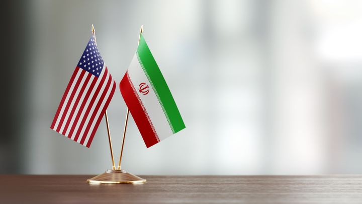 American and Iranian flag pair on desk over defocused background. Horizontal composition with copy space and selective focus.