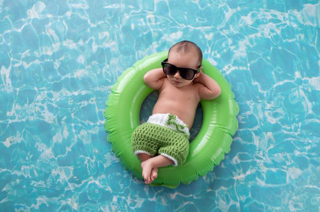 Two week old newborn baby boy sleeping on a tiny, green, inflatable swim ring. He is wearing green, crocheted board shorts and black sunglasses.